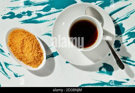 Eine weiße Tasse Kaffee, weiße Zuckerdose mit braunem Zucker und Löffel auf farbiger Baumwolltischdecke Stockfoto