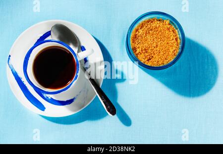 Eine Tasse Kaffee mit Löffel und braunem Zucker in blauer Zuckerdose, blauer Tischdecke Stockfoto