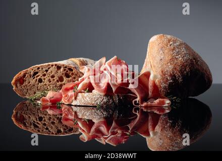 Prosciutto mit Ciabatta und Thymian auf schwarzem reflektierenden Hintergrund. Stockfoto