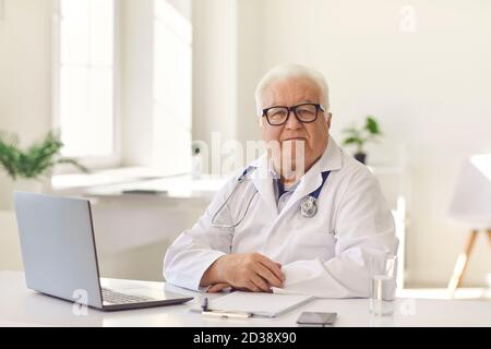 Serious Senior Arzt sitzt am Schreibtisch mit Laptop in modernen Krankenhausbüro mit Blick auf die Kamera Stockfoto