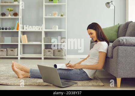 Lächelnder junger Student, der auf den Laptop-Bildschirm schaut und sich Notizen notiert Während des Online-Unterrichts Stockfoto