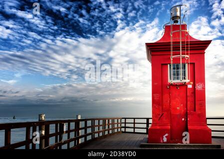 Kleiner roter Leuchtturm auf einem hölzernen Pier unter einem dramatischen Himmel. Lignano Sabbiadoro, Friaul Julisch Venetien, Italien. Stockfoto