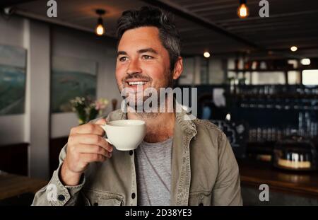 Hübscher weißer Mann lächelt, während er warmen Kaffee in schickem Café hält. Stockfoto