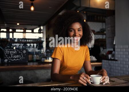 Schöne Frau mit afro sitzt im Café, lächelnd, während sie Keramik-Kaffeebecher. Stockfoto