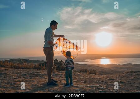Ein kleiner Junge und sein Vater spielen bei Sonnenuntergang fliegende Drachen in der Natur. Stockfoto