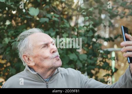 Ein älterer, aktiver, gutaussehender Mann mit Schnurrbart nimmt ein Selfie mit einem Handy. Ein älterer Mann schaut auf die Kamera und posiert scherzhaft Stockfoto