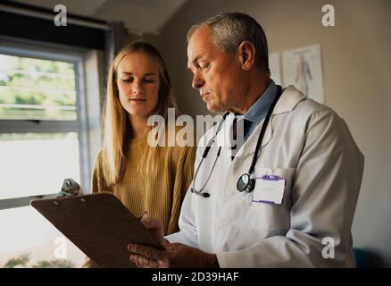 Ältere kaukasischen männlichen Arzt im Laborkittel Diagnose mit weiblichen Patienten diskutieren, Blick auf Zwischenablage Stockfoto