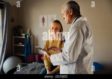 Männlicher Arzt, der mit weiblichen Patienten über Bluttestergebnisse spricht, sitzt auf ausklappbarem Bett im modernen Büro. Stockfoto