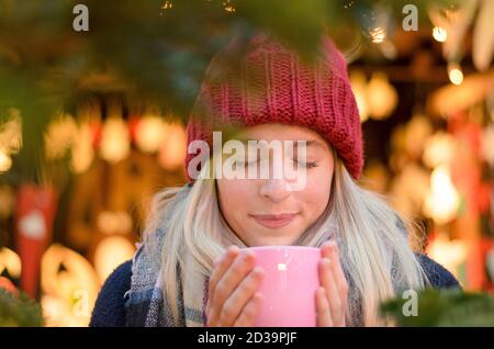 Freundliche junge Frau, die ein heißes Getränk genießen, wie sie besucht eine Straße Weihnachtsmarkt oder Messe umklammert eine große Tasse in den Händen gegen einen Bokeh bri Stockfoto