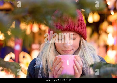 Freundliche junge Frau, die ein heißes Getränk genießen, wie sie besucht eine Straße Weihnachtsmarkt oder Messe umklammert eine große Tasse in den Händen gegen einen Bokeh bri Stockfoto