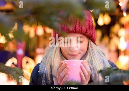 Freundliche junge Frau, die ein heißes Getränk genießen, wie sie besucht eine Straße Weihnachtsmarkt oder Messe umklammert eine große Tasse in den Händen gegen einen Bokeh bri Stockfoto