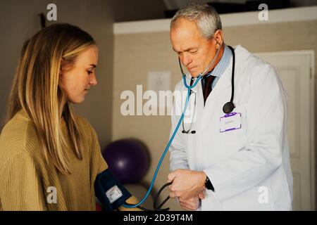 Ältere männliche Arzt sanft unter Blutdruck von jungen weiblichen Patienten sitzen in Arztpraxis. Stockfoto