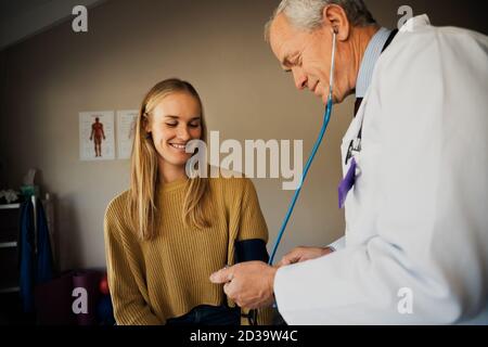 Ältere männliche Arzt nimmt Blutdruck von glücklichen weiblichen Patienten Stockfoto