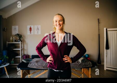 Blonde kaukasische weibliche Physio stehen mit Händen auf Hüften vor dem Bett in organisierten Studio. Stockfoto