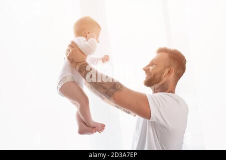 Fröhlich tätowierten Mann in weißem T-Shirt hält Säugling Sohn in Ausgestreckte Hände Stockfoto