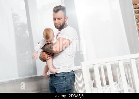 Tätowierte Mann hält Baby Junge, während in der Nähe von Krippe zu stehen Angezeigt Stockfoto