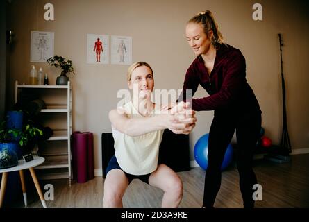 Junge weibliche Patientin üben hockende Position, während ausgebildete Physiotherapeut korrigiert Haltung in trendigen Studio Übung. Stockfoto