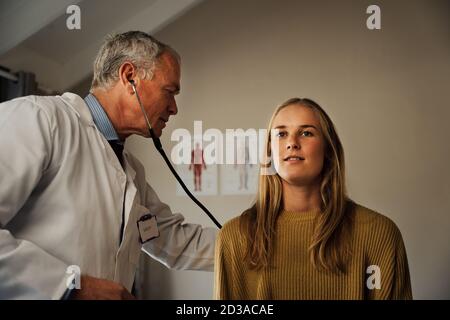 Älterer männlicher Arzt, der junge kaukasische Patientin mit Stethoskop untersucht Stockfoto