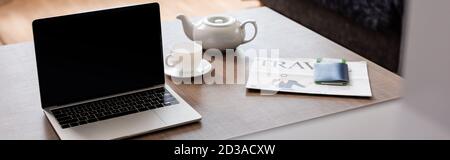 Panorama-Ernte von Laptop mit leerem Bildschirm in der Nähe Teekanne, Tasse, Reisezeitung und Pass mit Flugticket auf dem Tisch Stockfoto