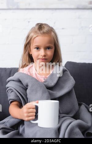 Selektiver Fokus von Kind in Decke gewickelt Blick auf die Kamera Während Sie die Tasse auf dem Tisch halten Stockfoto