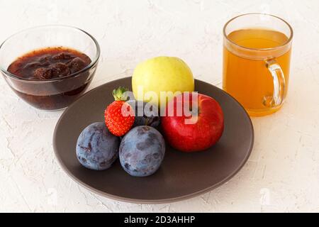 Gepflückt reife Pflaumen, Äpfel, eine Erdbeere auf dem Teller und eine Glasschale mit Marmelade, eine Tasse Tee auf dem weißen strukturierten Hintergrund. Nur geerntete Früchte Stockfoto