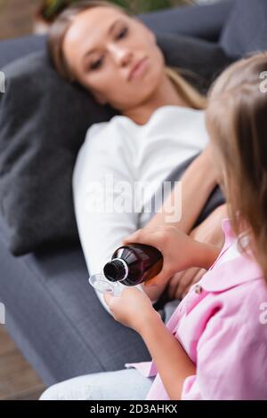 Selektiver Fokus von Kind Gießen Sirup in Löffel in der Nähe krank Mutter liegt auf der Couch Stockfoto