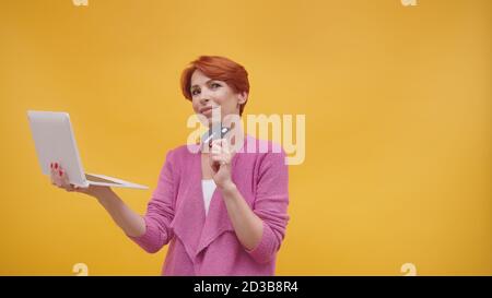 Rotschopf Frau mittleren Alters mit Laptop und Kreditkarte. Online-Shopping-Konzept. Isoliert auf orangefarbenem Hintergrund. Hochwertige Fotos Stockfoto