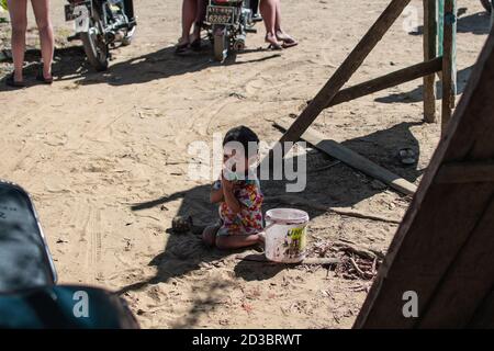 Ngwesaung, Myanmar - 26. Dezember 2019: Ein junges burmesisches Kind in einem bunten Kleid sitzt im Sand und hält ein Plastikspielzeug am 26. Dezember 2019 Stockfoto