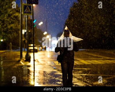 Blick auf eine Fußgängerüberführung in der Stadt bei Nacht bei starkem Regen. Silhouette eines Mannes mit einer Plastiktüte auf dem Kopf statt eines Regenschirms Stockfoto