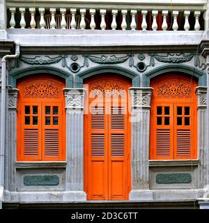 Restauriertes chinesisches peranakan-Ladenhaus mit orangefarbenen Lamellenfenstern aus Holz und verzierten Bögen und Architraven auf einer dunkelgrauen Steinwand. Stockfoto