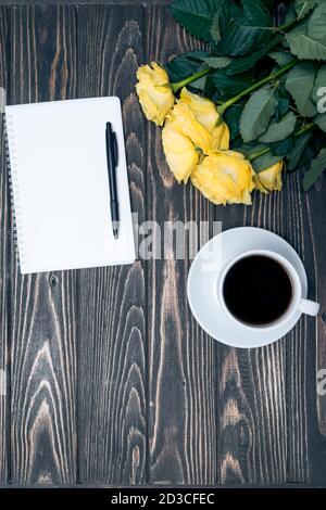 Notizbuch, Tasse Kaffee und gelbe Rose auf Holzhintergrund. Nachbildung Stockfoto