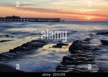 Sonne geht hinter Hastings Pier in Ost-Sussex mit dem Gezeiten am Strand von Saint Leonards enthüllen die Felsen Stockfoto