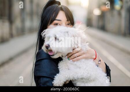 Horizontale Nahaufnahme Porträt der schönen Brünette Mädchen mit Pferdeschwanz Haar, umarmt ihren kleinen niedlichen Hund, posiert im Freien in alten europäischen Stadtstraße an Stockfoto