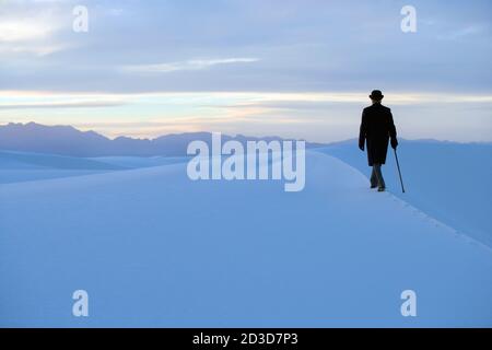 Rückansicht des Mannes in einem Mantel und Melone Hut, der durch eine weiße Dünenlandschaft mit einem Stock geht. Stockfoto