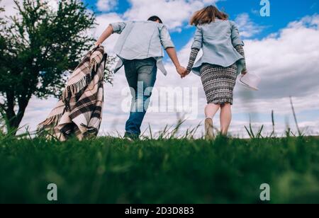 Junges Paar in der Liebe mit einem karierten karierten kariert läuft über ein grünes Feld halten Hände auf einem Hintergrund von blauem Himmel. Rückansicht und Unteransicht. Getöntes Bild Stockfoto