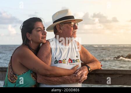 Eine Frau umarmt einen älteren Mann und ruht ihr Kinn während des Sonnenuntergangs am Meer auf seiner Schulter. Stockfoto