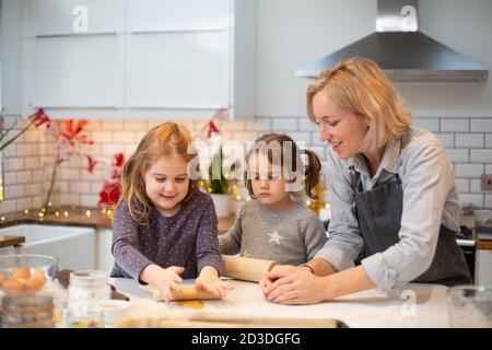 Blonde Frau trägt blaue Schürze und zwei Mädchen stehen in der Küche, Backen Weihnachtsgebäck. Stockfoto