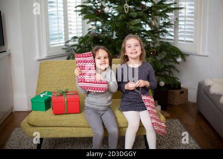 Zwei junge Mädchen sitzen auf dem Sofa im Wohnzimmer, halten rot-weißen Weihnachtsstrumpf und lächeln an der Kamera. Stockfoto