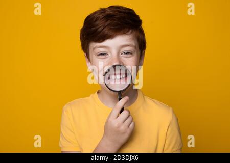 Ingwer kaukasischen Jungen hält eine Lupe auf seine Mund und Lächeln toothy auf Kamera posiert auf einem gelben studiowand Stockfoto