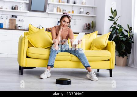 Frau sitzt auf der Couch mit digitalem Tablet in der Nähe von Roboter Vakuum Reiniger Stockfoto