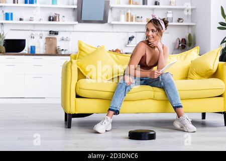 Frau schaut weg und hält das digitale Tablet in der Nähe des Roboter-Staubsaugers Stockfoto