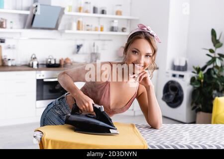 Junge Frau schaut auf die Kamera, während sie T-Shirt mit Dampf bügelt Zu Hause Stockfoto