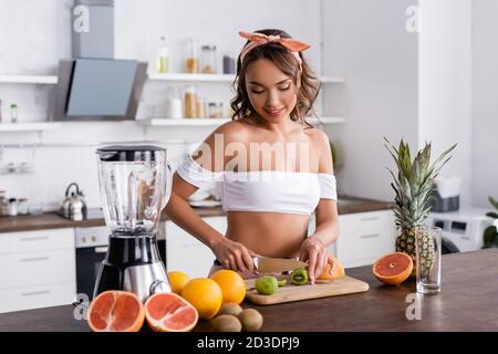 Selektiver Fokus der jungen Frau Schneiden Kiwi während der Zubereitung Smoothie In der Küche Stockfoto
