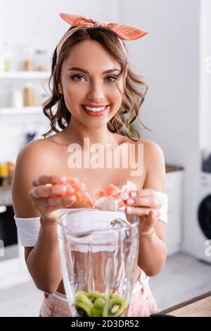 Selektiver Fokus der Hausfrau Blick auf die Kamera, während das Hinzufügen von Früchten Im Mixer Stockfoto