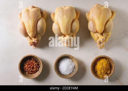 Drei rohe organische ungekocht ganz gelb Mais mini Huhn in der Zeile mit Salz, Kurkuma Pulver, red hot chili Pfeffer in Schalen über Marmor Hintergrund. Coo Stockfoto