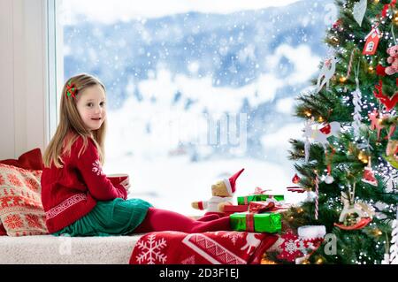 Kind trinken heiße Schokolade am Weihnachtsbaum zu Hause. Kid öffnen Geschenke und Geschenke. Kleines Mädchen in Santa kleid und hut mit heißem Kakao in Rot Stockfoto