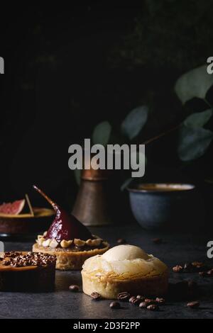 Vielzahl von süßen Törtchen mit Schokolade, Karamell, Birnen, Feigen mit Tasse Kaffee und Kaffeebohnen auf schwarze Textur Tabelle. Stockfoto