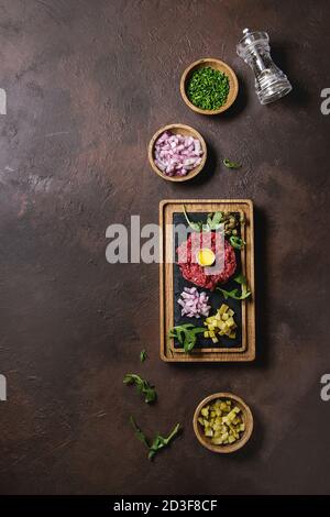 Beef Tartare mit Wachteleier in der Schale, schneiden Gewürzgurken, Kapern, rote Zwiebel, Schnittlauch, Rucola auf hölzernen schwarzer Schiefer mit Pfeffer mil serviert. Stockfoto