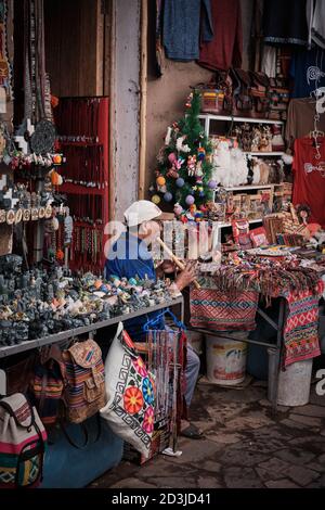 Pisaq Markt Pisac Kleinbauer spielt auf einer Blockflöte oder einem ähnlichen Holzblasinstrument, um Touristen anzuziehen. Er ist von bunten Geschenken umgeben Stockfoto