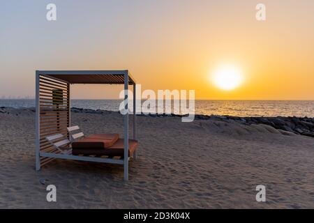 Leere Doppel-Sonnenliege am Sandstrand in Dubai, mit orangefarbenem Sonnenuntergang im Hintergrund und ruhigem Meer. Stockfoto